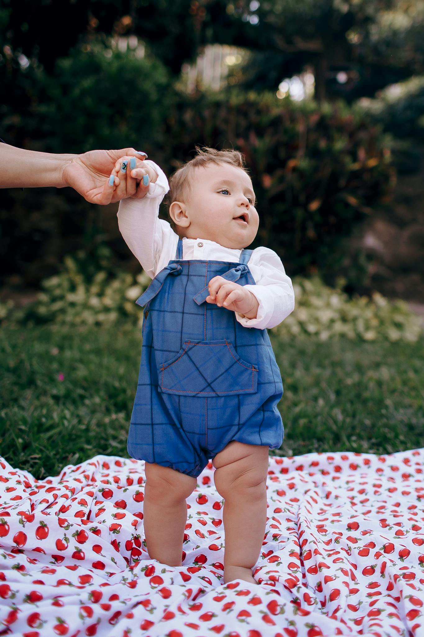 Charming baby in blue overalls holding a parent's hand, enjoying playtime outdoors on a summer day.